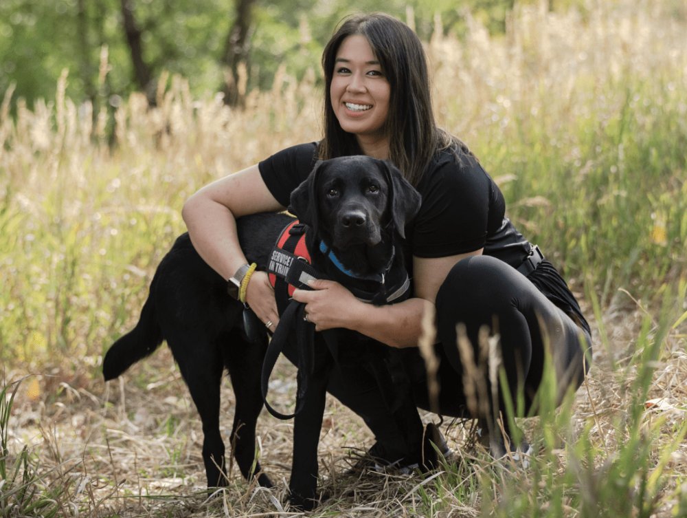 Lisa Gallegos crouching beside a black Labrador wearing a service-dog-in-training vest